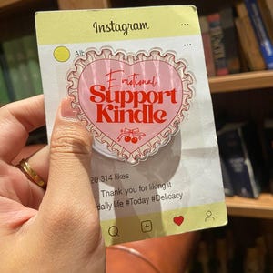 May include: A pop-up phone grip with a pink heart design that reads "Emotional Support Kindle" in red script. The grip is designed to look like an Instagram post, with the word "Instagram" at the top.