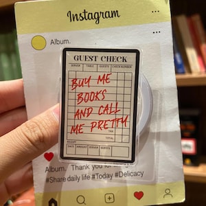 May include: A clear phone grip with a "Guest Check" design, featuring handwritten text that reads "Buy Me Books And Call Me Pretty." The grip is attached to a light yellow Instagram-themed card.