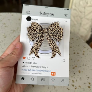 May include: A phone grip featuring a tan and black leopard print bow design, mounted on a rectangular display card styled like an Instagram post. The card includes the text Album, 520,1314 Likes, Thank you for liking it, and hashtags #Share daily life# #Today# #Delicacy#.
