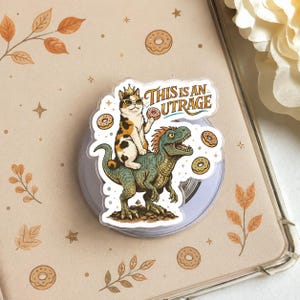 May include: A clear phone grip featuring a cat wearing sunglasses and a crown riding a green dinosaur, with the text "THIS IS AN UTRAGE". The cat and dinosaur are surrounded by donuts.