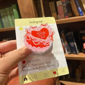 May include: A pink and red cake-shaped phone grip with the text "I'm literally just a girl" on a card designed to look like an Instagram post. The card has a yellow border and includes social media icons.