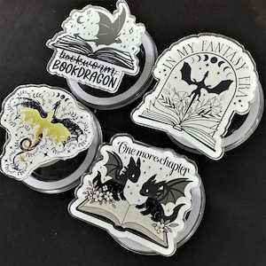 May include: Four clear acrylic phone grips with dragon and book designs. One says "bookworm BOOKDRAGON", another says "IN MY FANTASY ERA", and a third says "One more chapter". The fourth grip has a yellow dragon.
