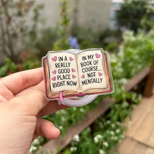 May include: A clear acrylic phone grip featuring a book design with the text "In a really good place right now" and "In my book of course... not mentally". The book has a pink ribbon bookmark and pink hearts.