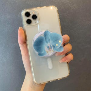 May include: A clear smartphone case featuring a triple-lens camera array and a circular phone grip attachment. The grip displays a detailed, high-resolution image of a blue orchid flower with white and yellow accents at its center.