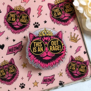 May include: A circular phone grip featuring a pink cat wearing a gold crown and round sunglasses with the text THIS IS AN OUT RAGE! printed on the lenses. The background is a light pink surface with a repeating pattern of the same cat, crowns, stars, lightning bolts, and the word MEOW! in speech bubbles.