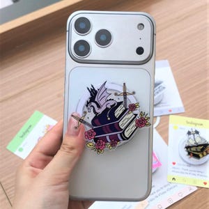 May include: A silver phone case with a fantasy-themed pop socket. The pop socket features a purple and white dragon, two crossed swords, and two stacked books with pink roses. The phone case is held by a hand.
