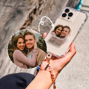 May include: A clear smartphone case featuring a custom photo of a couple attached to the back. The case includes a colorful beaded wrist strap with a small metal tag. A circular inset shows the original photograph of the couple used for the custom piece.