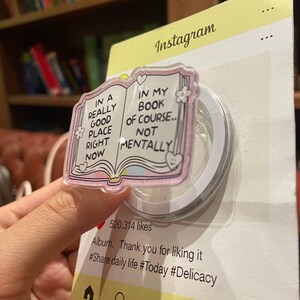 May include: A clear, iridescent phone grip with a pink book design and the text "IN A REALLY GOOD PLACE RIGHT NOW" and "IN MY BOOK OF COURSE.. NOT MENTALLY". The grip is attached to a yellow Instagram-themed backing.