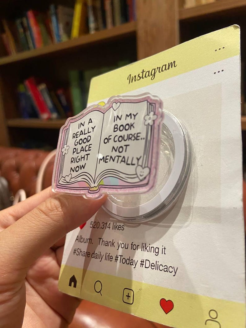 May include: A clear, iridescent phone grip with a pink book design and the text "IN A REALLY GOOD PLACE RIGHT NOW" and "IN MY BOOK OF COURSE.. NOT MENTALLY". The grip is attached to a yellow Instagram-themed backing.