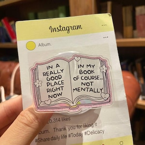 May include: A clear phone grip with a pink and glittery border, shaped like an open book. The book design has the text "IN A REALLY GOOD PLACE RIGHT NOW" and "IN MY BOOK OF COURSE.. NOT MENTALLY". The grip is on a yellow Instagram-themed card.