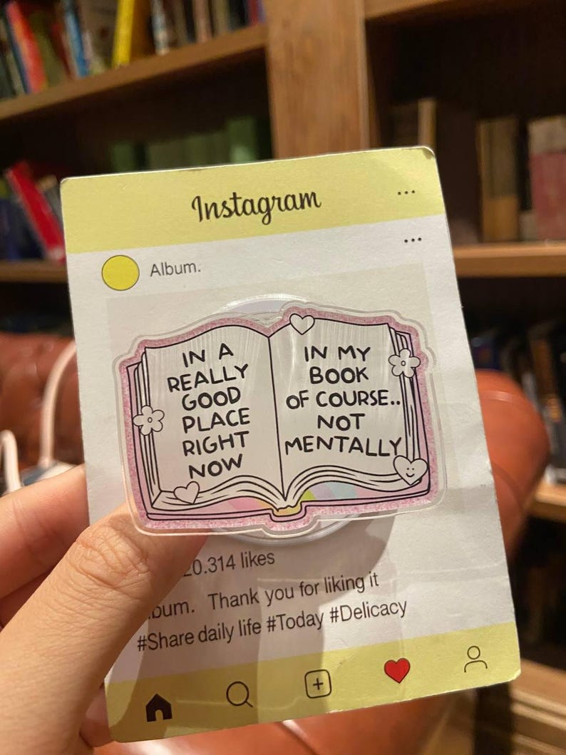 May include: A clear phone grip with a pink and glittery border, shaped like an open book. The book design has the text "IN A REALLY GOOD PLACE RIGHT NOW" and "IN MY BOOK OF COURSE.. NOT MENTALLY". The grip is on a yellow Instagram-themed card.