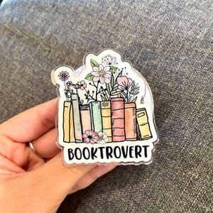 May include: A clear, die-cut sticker featuring a stack of books with colorful spines, topped with a variety of flowers and greenery. The word "BOOKTROVERT" is printed in black block letters at the bottom of the design.