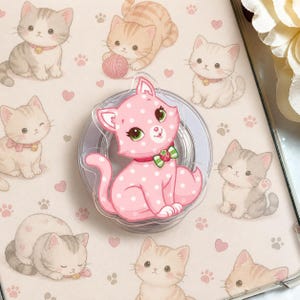 May include: A pink cat phone grip with white polka dots and a green bow tie, surrounded by illustrations of cartoon kittens in various poses. The background features paw prints and hearts on a light pink surface.