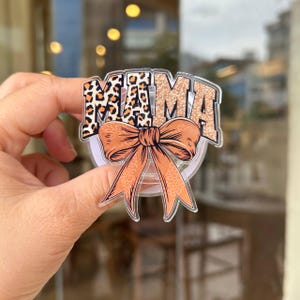 May include: A circular phone grip featuring the word MAMA in a bold, block font with a brown and black leopard print pattern. Below the text is a large, peach-colored bow with a subtle leopard print texture, all set on a clear acrylic base.