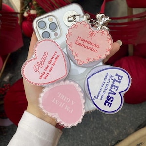 May include: A clear phone case with a silver keychain holding several heart-shaped charms. The charms have phrases like "Please skip the flowers buy me books" and "Hopeless Romantic" in pink and white.
