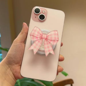 May include: A light pink smartphone featuring a dual-camera system and a circular phone grip attached to the back. The grip is adorned with a prominent, flat, acrylic-style decorative element shaped like a bow with a pink and white gingham pattern.