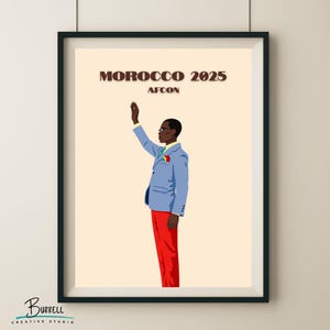 May include: A framed art print featuring a stylized illustration of a person in a blue suit and red pants, raising their hand. The text "MOROCCO 2025 AFCON" is above the figure. The artwork is set against a cream background.