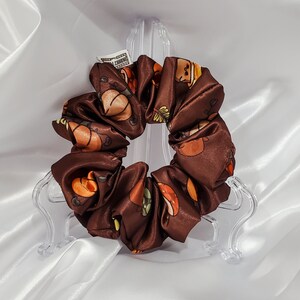 May include: A brown satin scrunchie with a repeating pattern of cartoon pumpkins and autumn leaves in orange, yellow, and green. The scrunchie is displayed on a clear plastic stand against a white satin background.