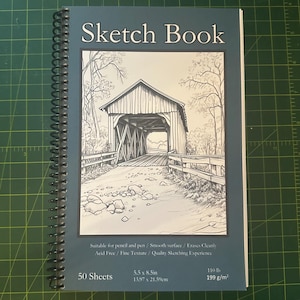 May include: A blue sketchbook with a black and white illustration of a covered bridge on the front. The cover reads "Sketch Book" and includes details about the paper: 50 sheets, 5.5 x 8.5 inches, 110 lb, 199 g/m².
