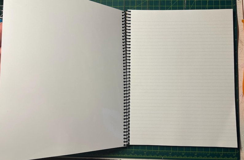 Puede incluir: Cuaderno abierto con espiral y p&aacute;ginas blancas en blanco. La p&aacute;gina izquierda est&aacute; vac&iacute;a, mientras que la p&aacute;gina derecha presenta l&iacute;neas horizontales. El cuaderno est&aacute; abierto sobre una esterilla de corte verde.