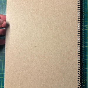 Puede incluir: Un cuaderno con espiral y cubierta de papel kraft. Las p&aacute;ginas est&aacute;n en blanco y la encuadernaci&oacute;n es negra. El cuaderno est&aacute; abierto, revelando las p&aacute;ginas interiores. El fondo es una esterilla de corte verde.