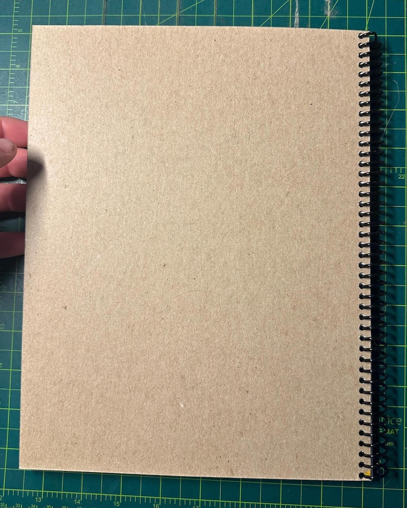 Puede incluir: Un cuaderno con espiral y cubierta de papel kraft. Las p&aacute;ginas est&aacute;n en blanco y la encuadernaci&oacute;n es negra. El cuaderno est&aacute; abierto, revelando las p&aacute;ginas interiores. El fondo es una esterilla de corte verde.