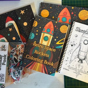 May include: A collection of spiral-bound notebooks and a sticker pack. The notebooks feature space-themed designs with rockets, planets, and stars on a black background. One notebook is a coloring book, and another is a sketchbook.