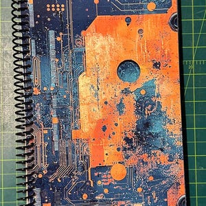 Puede incluir: Cuaderno con espiral con un dise&ntilde;o de placa de circuito. La cubierta presenta un patr&oacute;n abstracto azul y naranja que se asemeja a componentes electr&oacute;nicos. El cuaderno tiene una encuadernaci&oacute;n en espiral negra.
