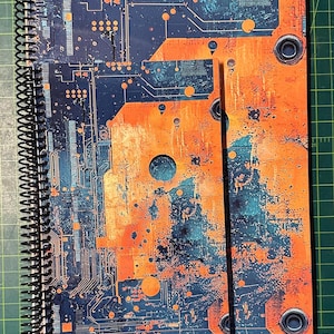 Puede incluir: Un cuaderno con espiral con un dise&ntilde;o vibrante de placa de circuito en tonos naranja, turquesa y azul marino. La cubierta muestra intrincados caminos y componentes electr&oacute;nicos, creando una est&eacute;tica inspirada en la tecnolog&iacute;a.