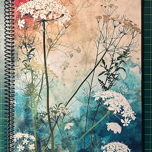 May include: Spiral-bound notebook featuring a botanical design. The cover art showcases white wildflowers and delicate stems against a backdrop of watercolor washes in shades of blue, orange, and beige. The notebook's spiral binding is black.