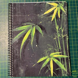 May include: Spiral-bound notebook with a black cover featuring a bamboo design. The cover has a dark background with green and yellow bamboo leaves and stalks. The notebook's spiral binding is black.