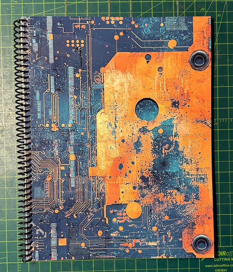 Puede incluir: Un cuaderno con espiral con un dise&ntilde;o de placa de circuito en tonos azules, naranjas y cobrizos. La cubierta tiene un aspecto envejecido y texturizado, con un gran panel naranja en el lado derecho. El cuaderno est&aacute; sobre una esterilla de corte verde.