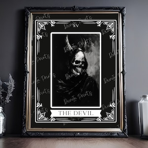 May include: Black framed art print featuring a dark illustration of a skull-like figure with horns, labeled "THE DEVIL." The artwork is set against a black background with decorative white borders. The print is displayed on a wooden surface.