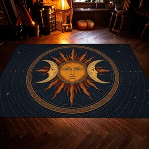 Puede incluir: Una alfombra rectangular con un diseño celestial. La alfombra tiene un fondo azul oscuro con un gran sol en el centro, rodeado de dos lunas crecientes. El sol y las lunas son en tonos dorados y naranjas. La alfombra está colocada sobre un suelo de madera.