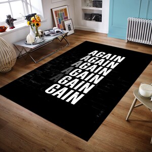 K&ouml;nnte beinhalten: Ein rechteckiger schwarzer Teppich mit dem Wort "AGAIN" in wei&szlig; vertikal wiederholt, mit einer grauen Instanz. Der Teppich hat eine ziegelartige Textur. Der Teppich liegt auf einem Holzboden.