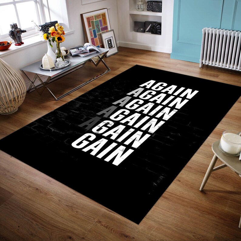 K&ouml;nnte beinhalten: Ein rechteckiger schwarzer Teppich mit dem Wort "AGAIN" in wei&szlig; vertikal wiederholt, mit einer grauen Instanz. Der Teppich hat eine ziegelartige Textur. Der Teppich liegt auf einem Holzboden.