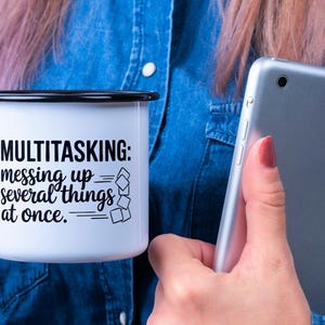 May include: White enamel mug with a black rim and handle. The mug has a black graphic that says "MULTITASKING: messing up several things at once." The graphic includes a stack of squares that are falling over.