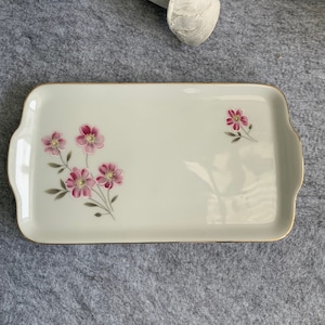 Könnte beinhalten: Ein rechteckiges Serviertablett in Weiß mit goldener Einfassung und einem Blumendesign mit rosa Blüten und grünen Blättern.