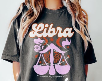 May include: A black t-shirt with a pink and orange Libra zodiac sign graphic. The graphic features a stylized scale with the text "I'm not indecisive, I'm just weighing my options."