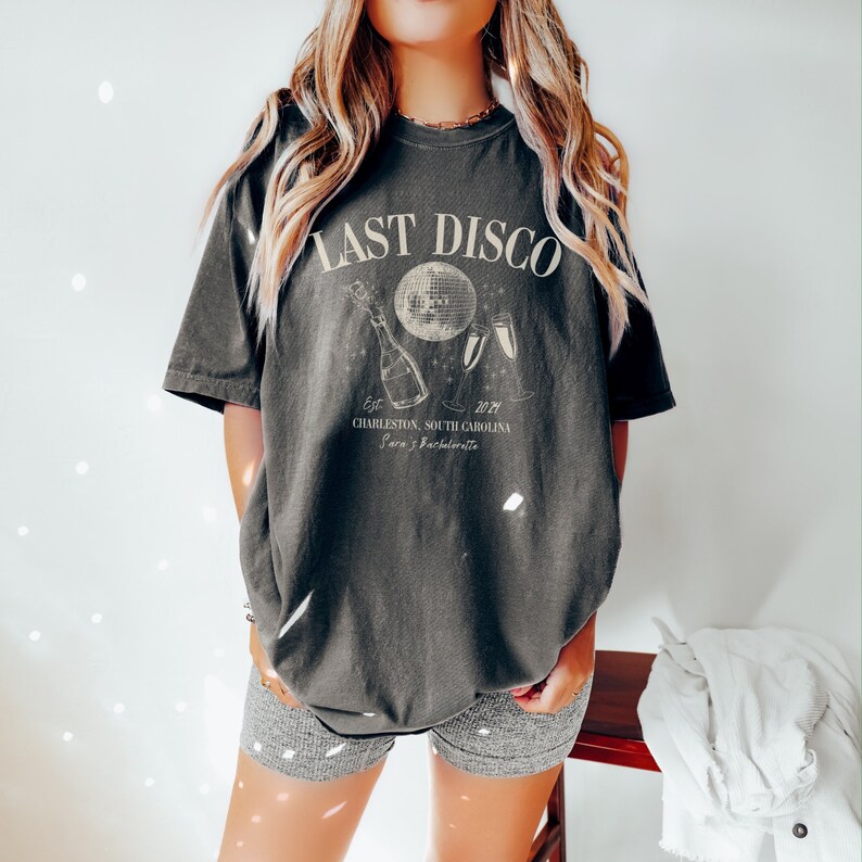May include: A dark gray t-shirt with the text "Last Disco" printed on the front. The shirt also features a disco ball, champagne bottle, and two champagne flutes. The text "Est. 2021 Charleston, South Carolina Sara's Bachelorette" is printed below the graphic.