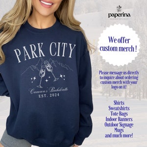 K&ouml;nnte beinhalten: Ein marineblauer Sweatshirt mit dem Text "PARK CITY" in wei&szlig;en Buchstaben. Der Sweatshirt zeigt eine Grafik von Bergen mit zwei Champagnergl&auml;sern, die ansto&szlig;en. Der Text "Cameron's Bachelorette EST. 2024" ist ebenfalls auf dem Sweatshirt aufgedruckt.