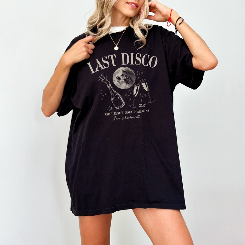May include: Black t-shirt with a white graphic design that says "Last Disco" with a disco ball, champagne bottle, and two champagne glasses. The text below the graphic says "Est. 2021 Charleston, South Carolina Sara's Bachelorette"