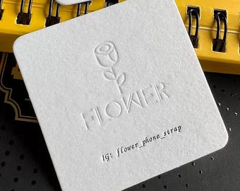 May include: Square white card with embossed flower design and the word "FLOWER". The text "IG: flower_phone_strap" is printed below. The card is on a dark surface with a yellow spiral notebook in the background.