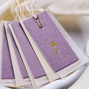 Pode incluir: Uma pilha de etiquetas cor de lavanda com texto dourado e um design floral. As etiquetas são presas com barbante branco e um fecho dourado. O texto na etiqueta diz "BEAUTY CANDY" e "I HAVE MY STYLE."