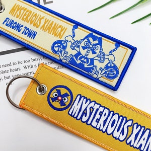 May include: Two yellow and blue keychains with the text "MYSTERIOUS XIANGXI" and a cartoon character. The top keychain has the text "FURONG TOWN" and the bottom keychain has a cartoon character with a red nose and blue eyes.