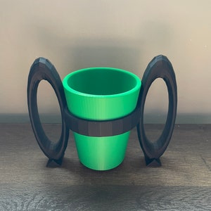 May include: A 3D printed black cup holder with two handles. The holder is designed to hold a green cup.