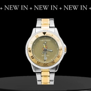 May include: A two-tone gold and silver wristwatch with a black face and a gold-colored Las Vegas Polo Club logo. The watch has a black bezel with white numbers and a date window at the 3 o'clock position.