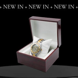 May include: A gold and silver wristwatch with a black face and white hands is displayed in a dark red wooden box with a white interior.