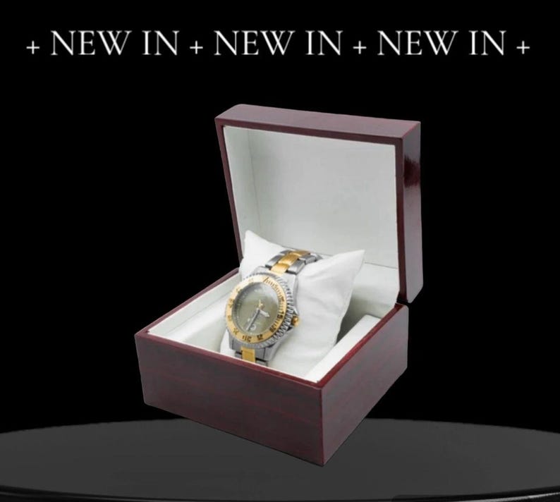 May include: A gold and silver wristwatch with a black face and white hands is displayed in a dark red wooden box with a white interior.