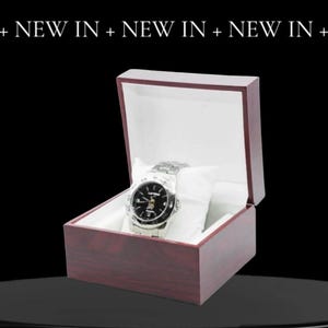 May include: A silver wristwatch with a black face and silver hands in a dark red wooden box with a white interior.